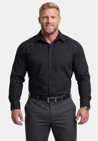 Smilende mand med kort blondt hår iført en sort skjorte med knapper, grå bukser, sort bælte og armbåndsur mod en ensfarvet baggrund.