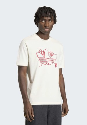 Muž s krátkymi dredmi, oblečený v bielej tričku s červeným abstraktným logom a tmavými nohavicami, stojaci na svetlom pozadí.