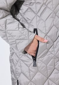 Grå quiltet jakke med diamantmønster, der har en sort lynlås-lomme og en blød, glat tekstur. Casual og funktionelt design.