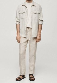 Licht beige linnen overhemd met twee voorzakken, gecombineerd met bijpassende broeken met een trekkoord in de taille. Zwarte sandalen maken de outfit compleet.