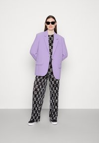 Blazer lavanda com dois bolsos frontais, combinado com calças largas com padrão preto e branco e ténis pretos. Óculos de sol redondos usados.