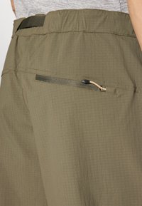 Calções verde-azeitona feitos de tecido texturizado, com um cós elástico e um bolso traseiro com fecho e uma pequena aba.