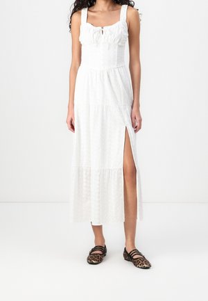 Witte maxi-jurk met een nauwsluitend lijfje, kanten details en een hoge split aan de dij; gedragen met bruine ballerina's met luipaardprint. Soepele textuur, zonder patronen.