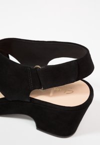 Sandale compensée en daim noir avec bride réglable à la cheville et semelle intérieure beige matelassée, marquée de lettres dorées.