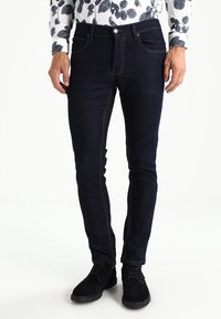 Tmavě modré džíny se slim-fit střihem, přední kapsy a kontrastní šití. V kombinaci s černými botami, vystavenými na neutrálním pozadí.