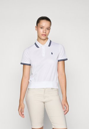 White polo shirt with navy-striped collar and cuffs, short sleeves, and a logo on the chest. Paired with beige shorts.