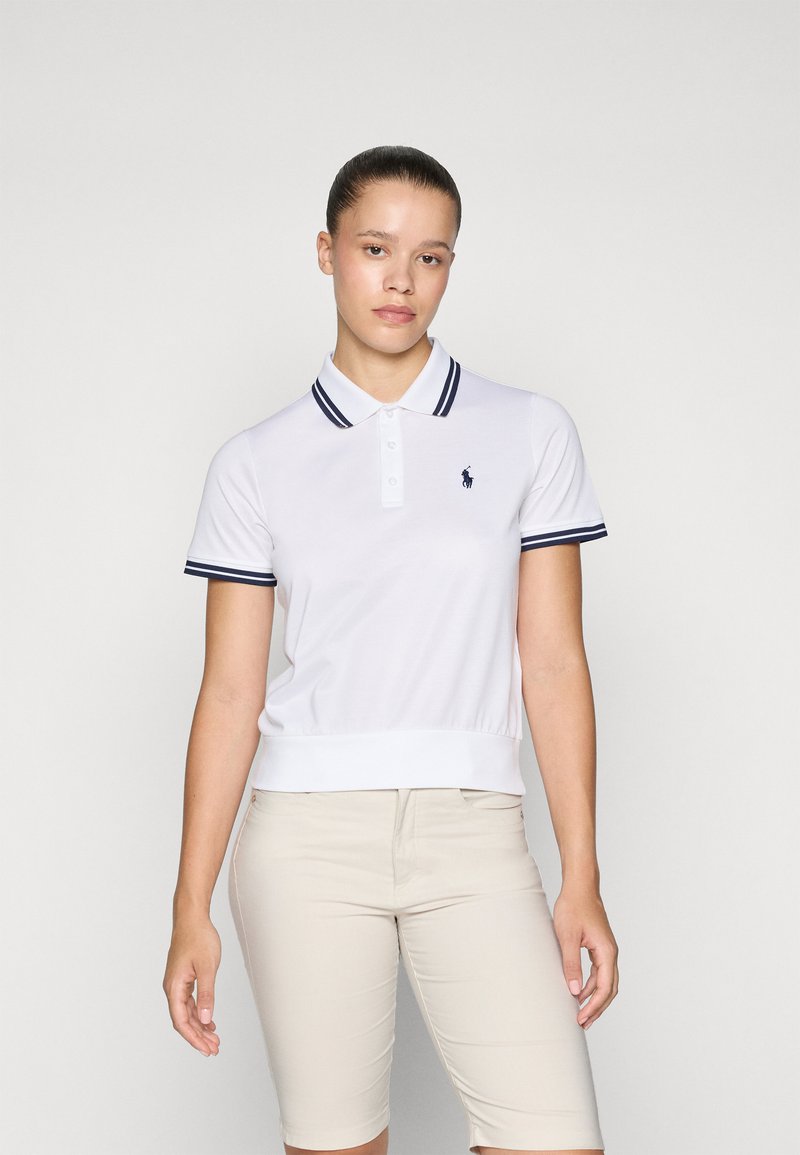 White polo shirt with navy-striped collar and cuffs, short sleeves, and a logo on the chest. Paired with beige shorts.