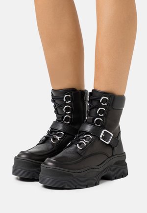 Bottines noires en cuir texturé, dotées de ferrures argentées, d'un laçage, d'une sangle ajustable et d'une semelle épaisse en caoutchouc.