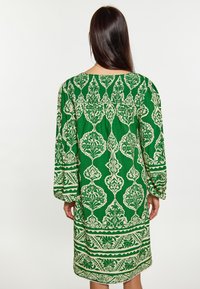 Rochie verde cu model, cu mâneci lungi, cu desene florale și geometrice albe complexe, cu un croi lejer și un tiv care ajunge până la genunchi.