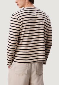 Osoba v béžovém a černém pruhovaném tričku s dlouhým rukávem a béžových kalhotách, s rukama v kapsách, otočená zády k jednobarevnému pozadí.