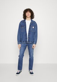 Chaqueta de denim con costuras más claras, dos bolsillos en el pecho y un parche con logo, combinada con jeans azules y zapatillas multicolores.