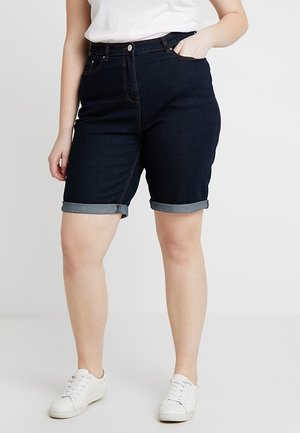 Personne portant un short en denim foncé à ourlet roulé, associé à un t-shirt blanc et des baskets blanches, debout devant un fond clair uni.