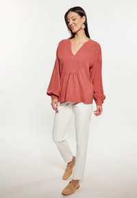 Blusa di colore ruggine, ampia e comoda, con scollatura a V, maniche a palloncino e dettaglio arricciato sotto il busto; abbinata a pantaloni bianchi e scarpe chiare.