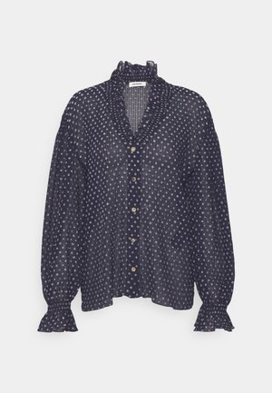 Frau trägt eine schwarze gemusterte Bluse mit Rüschenmanschetten, graue weit geschnittene Hose mit Kordelzug in der Taille und weiße Sneakers und steht drinnen.