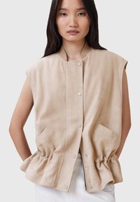 Gilet senza maniche in suede beige con un pannello frontale a coste, bottoni a pressione e vita elastica. Presenta tasche laterali e una silhouette rilassata.
