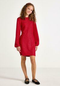 Rotes Glitzerkleid mit langen Ballonärmeln und geradem Schnitt. Verfügt über einen runden Ausschnitt und seitliche Schlitze. Kombiniert mit schwarzen flachen Schuhen.