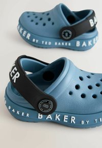 Modré plastové cлынky s perforáciami, zakriveným dizajnom a čiernym nastaviteľným popruhom s textom a logom "BAKER" v bielej farbe.