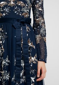 Marindräkt med långa genomskinliga ärmar, prydd med silver- och bronzfärgade paljetter i blommönster, med ett matchande satinbälte i midjan.