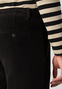 Črne kordrijeve hlače z vertikalno rebrasto teksturo, ki imajo zadaj žep s posameznim gumbom za zapiranje, prikazane ob rokavu črtaste srajce.
