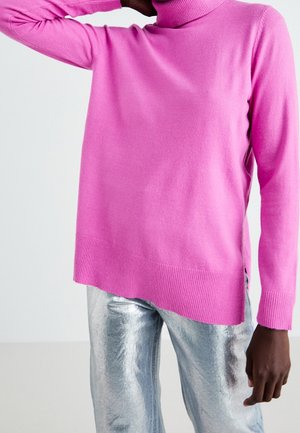 Personne portant un pull à col roulé rose vif associé à un pantalon métallique argenté brillant, avec un bras levé derrière la tête.
