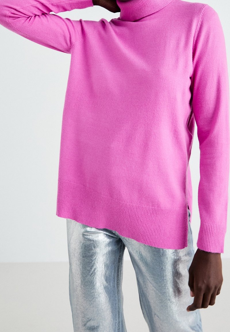 Personne portant un pull à col roulé rose vif associé à un pantalon métallique argenté brillant, avec un bras levé derrière la tête.