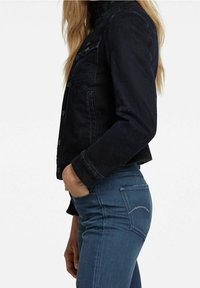 Tummansininen farkkutakki, jossa istuva leikkaus, napitettava edessä, rintatasku ja käännetty kaulus, yhdistetty sinisiin farkkuihin.