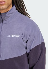 Fleece pullover i lys lilla og mørk lilla, med høj krave, halv lynlås lukning, og "TERREX" logo i hvidt på brystet.