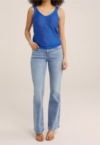 Blå ärmlös stickad topp med V-urringning, kombinerad med ljusblå utställda jeans. Skorna är beige sandaler med tunna remmar.