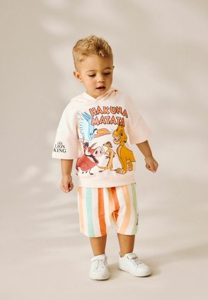 Hanorac roz deschis cu imprimeu "HAKUNA MATATA" care prezintă personaje, asortat cu pantaloni scurți multicolora cu dungi în nuanțe de portocaliu, verde și alb. Tenisi albi.