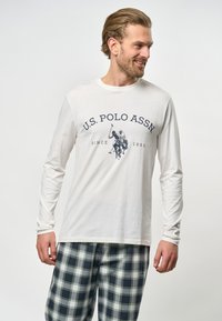 Hosszú ujjú fehér póló, tengerészkék nyomattal, amelyen az "U.S. POLO ASSN" felirat és egy pólós játékos grafika található, kockás tengerészkék és szürke nadrággal párosítva.