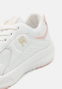 Valged tennisjalanõud, mis on valmistatud võrkmaterjalist ja nahast, heleda roosa aktsentidega, perforatsioonidega detailidega ning kuldse logoga küljel. Paks valge sisetald.