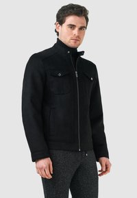 Svart mockajacka med dragkedja, två bröstfickor och hög krage. Texturerad yta med minimal hårdvara. Modellen bär mörkgrå byxor.