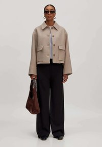 Vrouw in een beige knopenjasje, zwarte wijde broek, zwarte schoenen, zonnebril, hoepelringen, die een bruine handtas vasthoudt tegen een effen achtergrond.