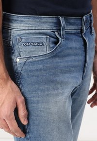 Близък план на човек, носещ светлосини дънки с видима етикет "Tom Tailor" на малкия преден джоб.