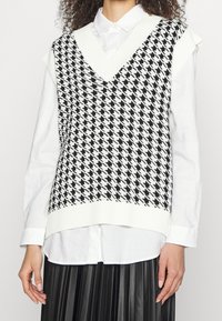 Veste sans manches en maille à motif pied-de-poule noir et blanc avec un décolleté en V profond, superposée sur une chemise blanche boutonnée. Détail à l'ourlet côtelé.