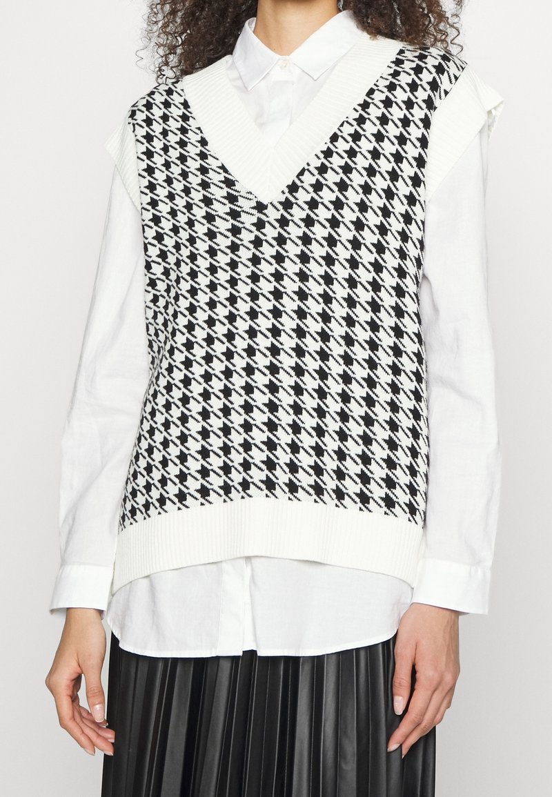 Veste sans manches en maille à motif pied-de-poule noir et blanc avec un décolleté en V profond, superposée sur une chemise blanche boutonnée. Détail à l'ourlet côtelé.