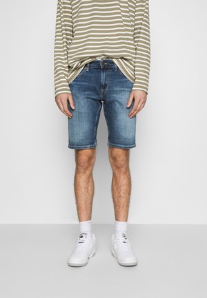 Mężczyzna w niebieskich szortach dżinsowych, białych sneakerach, białych skarpetkach oraz beżowo-białej koszuli w paski z długim rękawem, stojący na jednolitym tle.