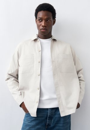 Young man wearing a light beige button-up shirt over a white sweatshirt and blue jeans, standing with hands in pockets against a white wall.