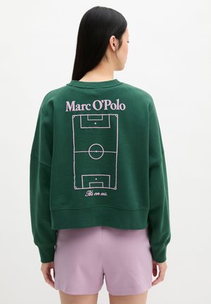 Femme aux longs cheveux foncés portant un sweat-shirt vert affichant un terrain de football et le texte "Marc O’Polo" dans le dos, associé à un short violet clair.