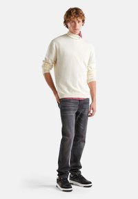 Pull à col roulé couleur crème, avec des poignets côtelés, associé à un jean gris foncé et des baskets noires à semelles blanches et lacets plats.