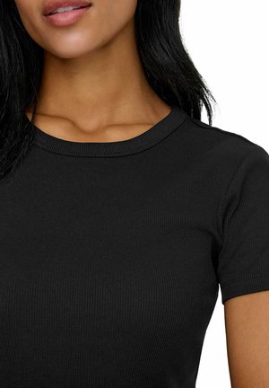 Frau trägt ein eng anliegendes schwarzes, geripptes Kurzarmshirt, das den Ausschnitt und einen Teil des Gesichts mit langen dunklen Haaren zeigt.