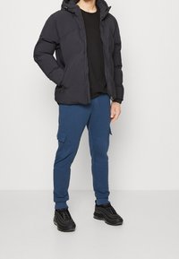 Man som bär en svart huva pufferjacka, svart t-shirt, blå cargojoggers och svarta sneakers, som står mot en enkel ljus bakgrund.