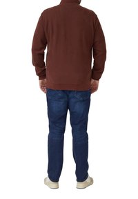 Brązowa teksturowana kurtka z wysokim kołnierzem, noszona z niebieskimi dżinsami i białymi trampkami, widziana od tyłu.