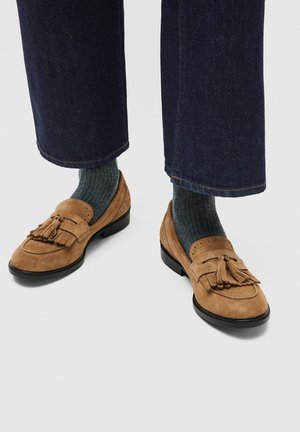 Pieds portant des chaussettes côtelées gris foncé, un jean court bleu marine et des mocassins à pampilles en daim beige avec semelles noires sur un fond clair.