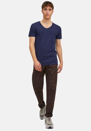 Ung mand går fremad iført en marineblå V-hals T-shirt, brune bukser med optrukne manchetter og grå sneakers mod en ensfarvet baggrund.