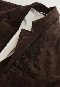 Hnědý manšestrový blazer se strukturovaným límcem, obsahující jeden kapsu a hladké béžové podšívání. Texturovaná látka s vertikálními žebry.