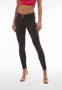 Femme portant un pantalon skinny taille haute noir, un top court rouge et des sandales à talons transparents, debout devant un fond blanc.