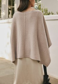 Lyserød uld poncho med en bred form, nedslået skulder og en glat tekstur, parret med en cremet nederdel med blonde detaljer.