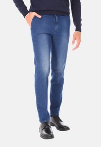 Jeans in denim blu slim-fit con una texture liscia, caratterizzati da un design a cinque tasche e dettagli di scoloritura delicati, abbinati a calzature nere.