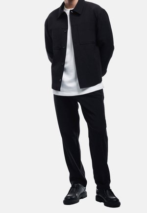 Mann trägt schwarze Jacke mit Knöpfen, weißes T-Shirt, schwarze Hose und schwarze Schuhe, steht mit den Händen hinter dem Rücken vor weißem Hintergrund.